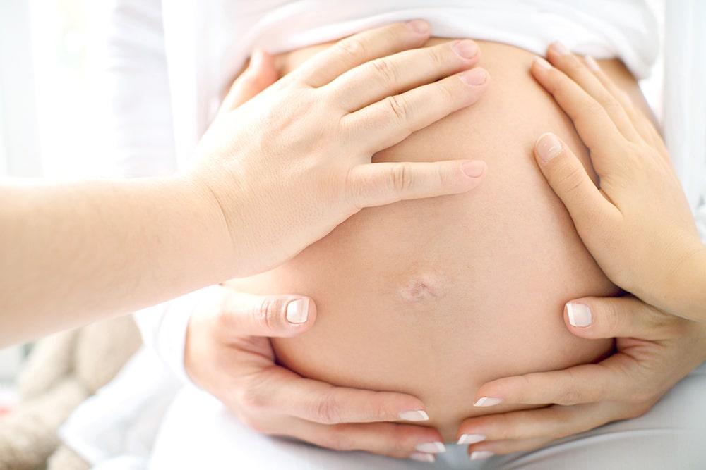 Several hands touch the belly of a pregnant woman. She hugs her stomach with well-groomed hands with a neat French manicure
