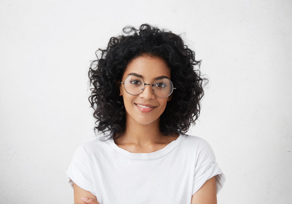 Close up studio shot of beautiful young mixed race woman model with curly dark hair looking at camera with charming cute smile while posing against white blank copy space wall for your content