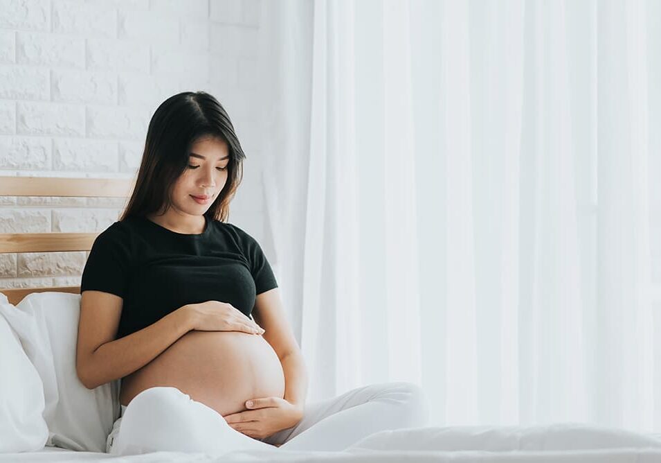 Happy Asian pregnant woman touching her belly with care in bedroom. She canât wait to be mom.